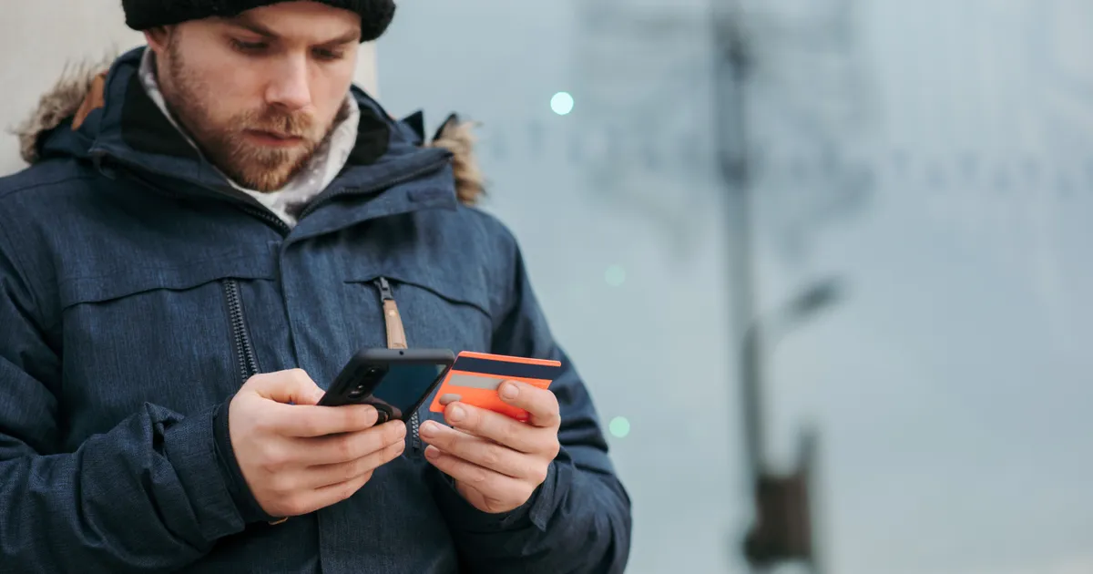 Man in a winter parka on a city street checking his banking app with his card out before applying for a $100 payday loan in Canada