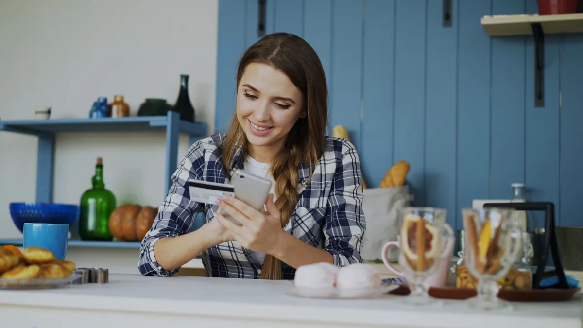 Woman in her kitchen reading her phone with her bank card in hand, checking what a $100 payday loan would leave from her paycheque