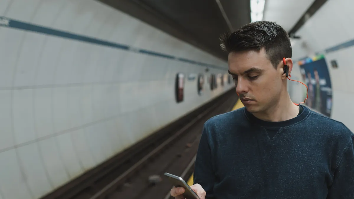 Young man on a subway platform reading his phone, working out whether a $100 payday loan or a pay advance covers him to Friday